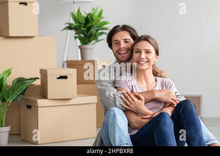 Cheerful excited millennial caucasian male and female in casual hugs, enjoy moving at new flat, sits among cardboard boxes with belongings in room. Bu Stock Photo