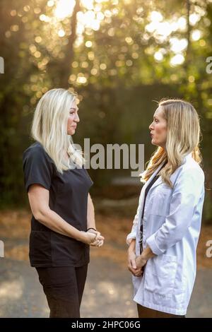 Two female medical professionals in black scrubs and one is wearing a white lab coat standing outside talking Stock Photo