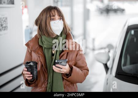 Woman in face mask holding smart phone and coffee cup while refueling car at gas station. Stock Photo