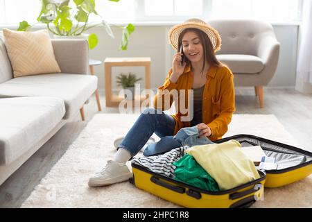Cool woman sitting near suitcase, speaking on cellphone, booking travel tour, making hotel reservation or buying tickets Stock Photo