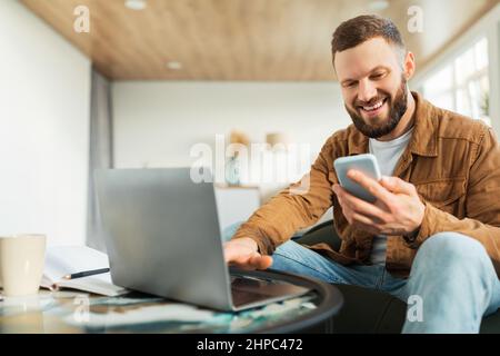 Male Entrepreneur Using Smartphone And Laptop Working Online At Home Stock Photo