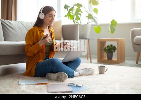 Young woman sitting on floor with laptop and headphones, studying online, having remote job, video conferencing at home Stock Photo