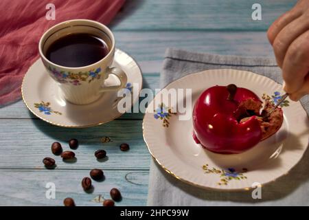 Breakfast concept with сherry cake on a ceramic plate, coffee in a faience cup, scattered coffee beans and a human hand taking a cake. Stock Photo