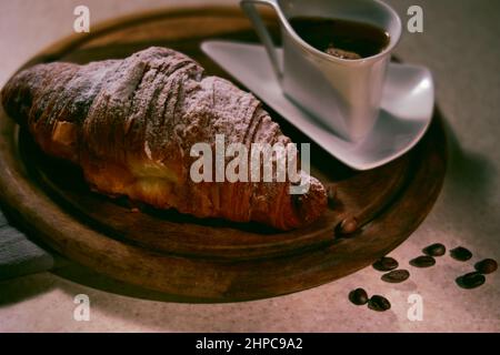 Breakfast concept with сroissant, coffee in a white faience cup on a wooden tray and scattered coffee beans on the table. Stock Photo