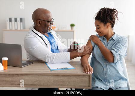 Vaccination In Pregnancy. Smiling Pregnant Young Black Woman Getting Vaccinated At Clinic, Mature Male Doctor In Glasses Giving Vaccine Shot Injecting Stock Photo