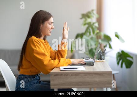 Online communication concept. Side view of millennial woman using laptop, waving at webcam, having video chat at home, free space. Beautiful Caucasian Stock Photo