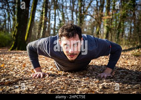 Runner is doing push-ups in the woods. The young man with the blue eyes and mustache is doing push ups on the ground while he listens to the music. Stock Photo