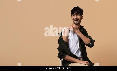 Cheerful middle eastern millennial man wearing eyeglasses with medical band on his shoulder looking at copy space and smiling over beige studio backgr Stock Photo