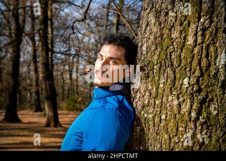 Portrait of a beautiful young man with blue eyes. The man is training in the woods and is leaning against an oak tree. Stock Photo