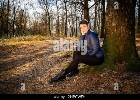 Young woman is sitting at the foot of an oak tree in the woods. Beautiful woman is resting after a walk in the park. Travel and outdoors concept. Stock Photo
