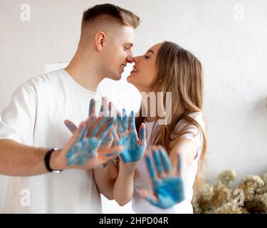Smiling young pretty couple with fair hair stand closing eyes, nuzzling noses, showing hands covered with blue paint. Stock Photo