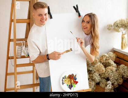 Happy young attractive couple stand behind easel with blank canvas and watercolor palette, posing, holding brushes.  Stock Photo