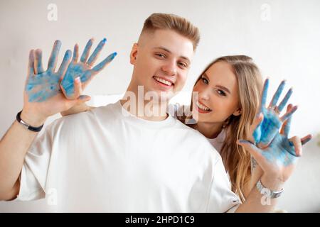 Happy smiling young couple of beautiful woman stand behind man in white T-shirts showing hands covered with blue paint. Stock Photo