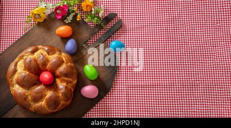 Easter Greek tsoureki braid top view. Sweet bread brioche and colorful egg on old rustic wooden baker shovel, checked red tablecloth Stock Photo