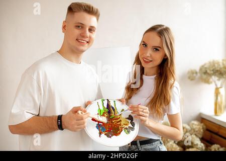 Portrait of happy smiling young pretty couple stand closely, posing, holding watercolor palette and brushes. Art therapy Stock Photo