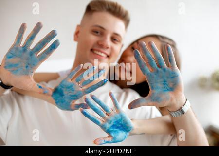 Happy smiling young couple of pretty woman stand behind man in white T-shirts showing hands covered with blue paint. Stock Photo