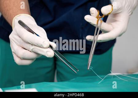 Doctor surgeon with disposable glove on hand holding forceps and scissor, stitch up wound or incision with suture thread over green fabric. Medical eq Stock Photo