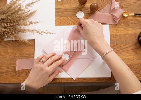 Cropped photo woman hands making greeting envelope craft, decorating with pink ribbon and stamping. Cereals on desk.  Stock Photo