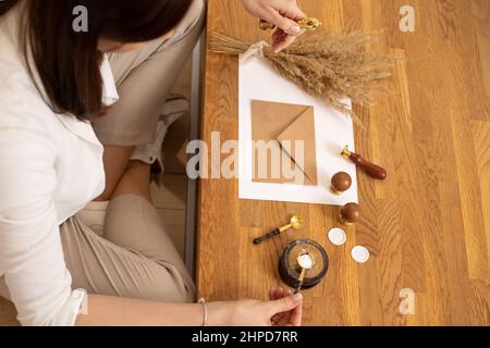 Cropped photo of woman hands making certificate present envelope craft, sitting at wood desk, holding paper, seal wax. Stock Photo