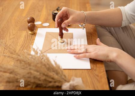 Cropped photo of woman hands making weeding invitation envelope craft, sitting at wood desk, putting sealing wax print.  Stock Photo