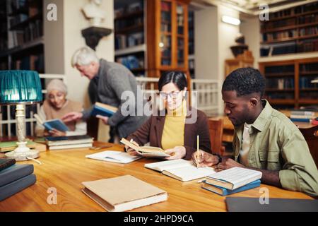 Diverse group of people sitting in row at table in college library and studying, focus on young black man reading book in foreground Stock Photo