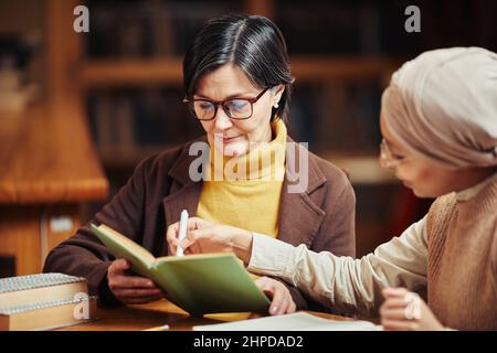 Warm toned portrait of two female adult students reading books while preparing for exam in college library Stock Photo