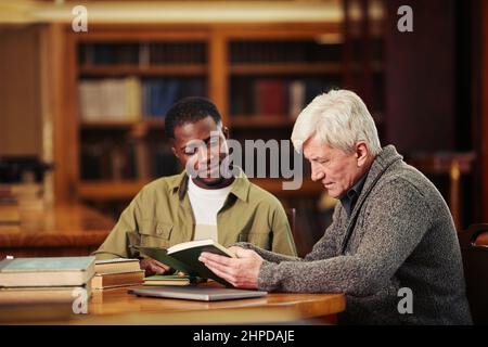 Portrait of male senior student reading book in library while preparing for exam with African-American friend Stock Photo