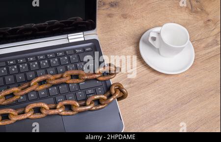 Computer keyboard locked by old rusty chain with reflection on display. Laptop detail and empty coffee cup on wooden desk. Bans and threats to freedom. Stock Photo