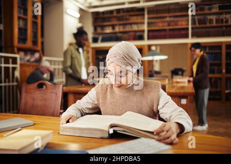 Warm toned portrait of young Muslim woman reading book in classic college library, copy space Stock Photo