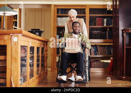 Full length portrait of smiling young man using wheelchair in college library with female friend assisting, copy space Stock Photo