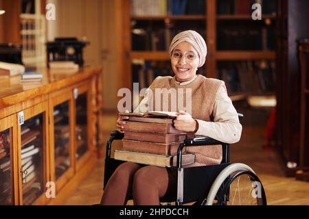 Warm toned portrait of smiling young woman using wheelchair in college library and looking at camera Stock Photo