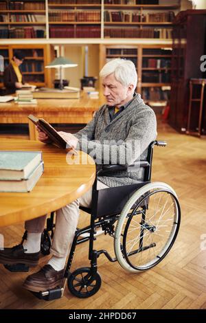 Vertical full length portrait of senior man using wheelchair and reading book in library Stock Photo