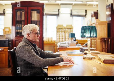Side view portrait of senior man with vision impairment reading book in braille at table in library Stock Photo