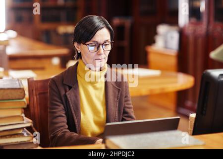 Warm toned portrait of female librarian using laptop while working with book catalogue Stock Photo