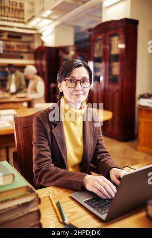Vertical portrait of female librarian using laptop while working with book catalogue Stock Photo