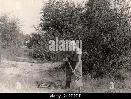 Trekhizbenka village, Lugansk region, USSR - circa 1982: Vintage photo of a soldier in a gas mask without a tunic. Humor. The man is holding a shovel. Stock Photo