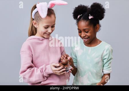 Waist up portrait of two happy girls eating chocolate eggs on Easter while standing against minimal background in studio Stock Photo