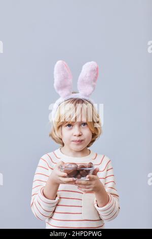 Waist up portrait of blonde boy holding chocolate eggs on Easter while standing against minimal background in studio Stock Photo