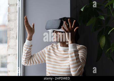 Stunned amazed young african woman gesturing in excitement while using VR headset glasses at home Stock Photo