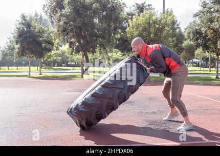 Bald man lifting and pushing a huge wheel exercising outdoors in a park Stock Photo