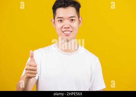 Asian handsome young man smiling positive showing thumbs up gesture good or like sign looking to camera, studio shot isolated on yellow background Stock Photo