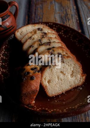 A clay or mud Mexican hand made plate, with sweet bread and raisins on top and a steaming cup of coffee or hot chocolate cocoa in the background. Stock Photo