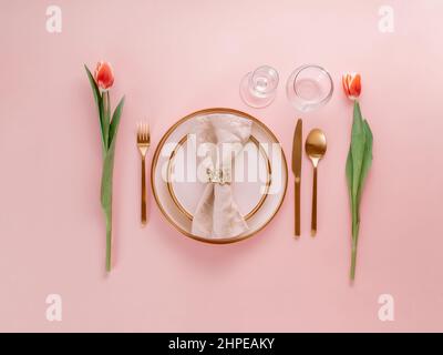 Festive pink table setting. Pink plate with gold cutlery and red tulip on pink background. Aesthetic stylish dinner table setting for Women's day, Mother's Day, wedding or anniversary. Flatlay topview Stock Photo