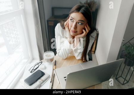 Beautiful woman sitting near bright window while  thinking about job  and holding her head. Portrait of female working with laptop computer on table Stock Photo