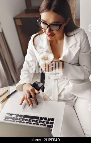 Beautiful young woman working on laptop computer while sitting at the living room, drinking coffee Stock Photo