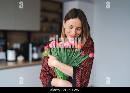Happy woman enjoy bouquet of tulips. Housewife enjoying a bunch of flowers and interior of kitchen. Sweet home. Allergy free Stock Photo