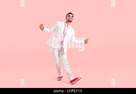 Happy funny young man in white suit, sneakers and glasses dancing on pink background Stock Photo
