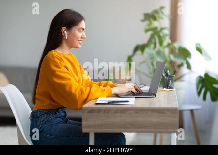 Cheery young woman in earphones using laptop, having online business meeting or educational webinar at home Stock Photo