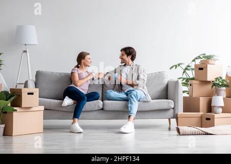 Glad young european couple in casual relaxing on sofa, drinking coffee, talking and planning new interior in room Stock Photo