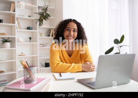Portrait of happy young black woman sitting at desk with study materials and laptop computer, smiling at camera indoors Stock Photo